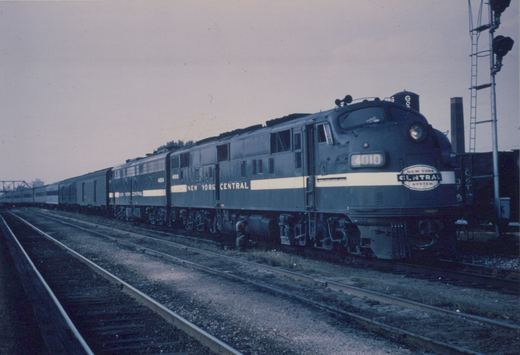 Two dark gray New York Central diesel passenger locomotives with a white side stripe, 4010 leading with its headlight on, pull a long passenger train past empty tracks under an overcast sky.
