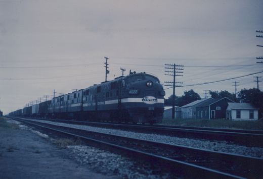 A dark diesel locomotive with a light stripe and an oval herald on its nose, its headlight lit, leads more units and a long line of boxcars past a pole line and a row of small frame buildings under a gray sky.