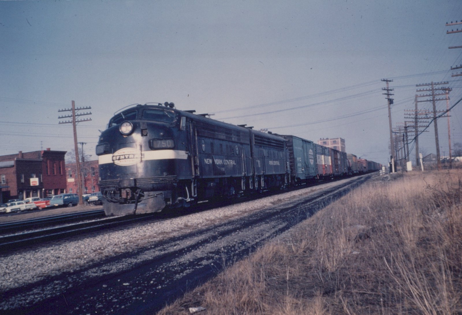 A black diesel locomotive with a white nose stripe and an oval New York Central herald leads a second unit and a long line of boxcars along a main line, with brick buildings and parked cars at left and dry grass and telegraph poles at right.
