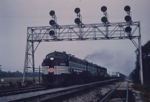 A dark New York Central diesel locomotive with white striped nose and lit headlight leads two more units and a freight train under a steel signal bridge with searchlight signals on an overcast day.