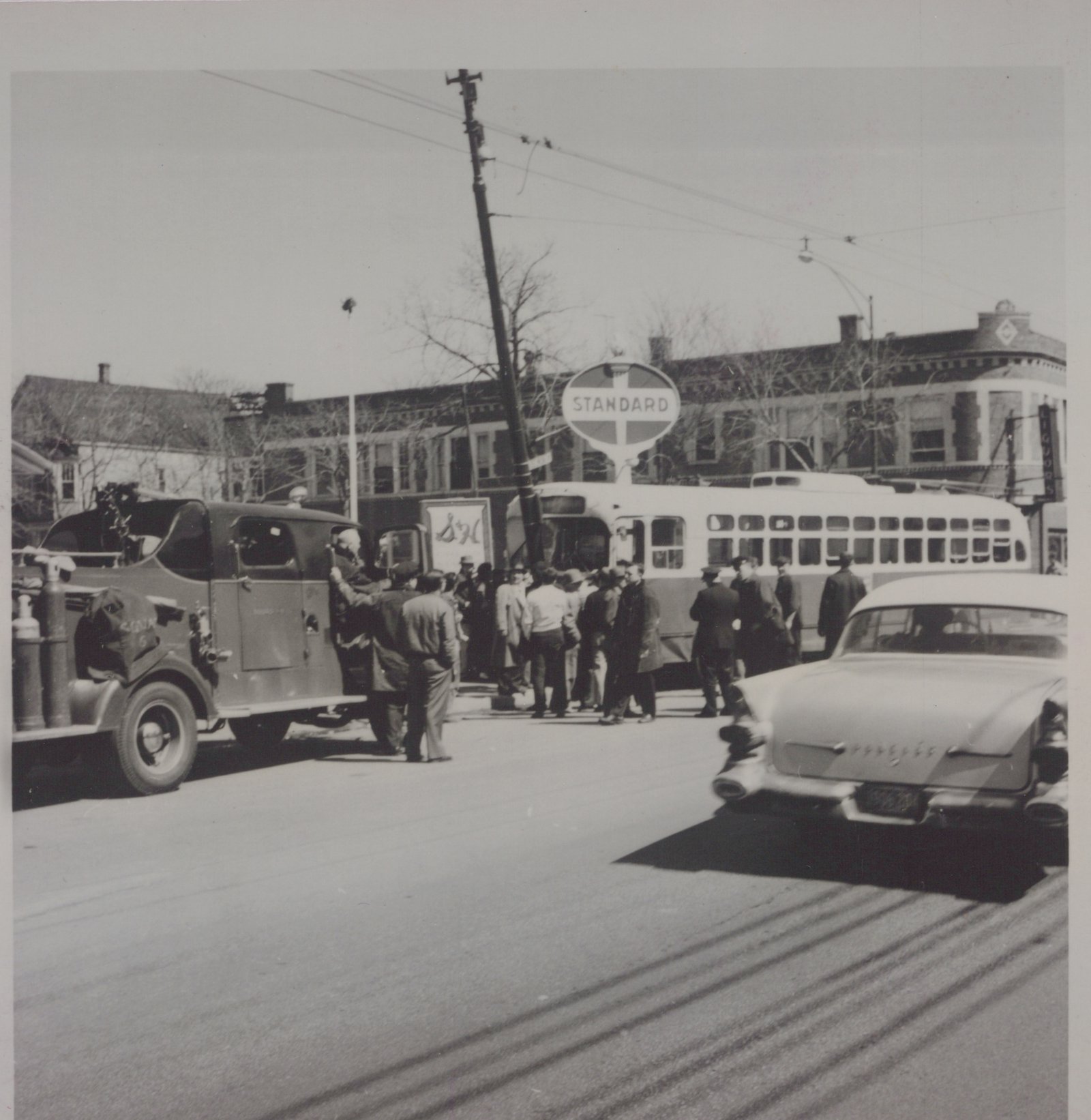 A black and white photograph of a street scene with a CTA PCC streetcar, a bus, a C&NW truck, and a Pontiac car, surrounded by people on a sunny day.