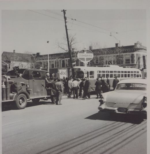 A black and white photograph of a street scene with a CTA PCC streetcar, a bus, a C&NW truck, and a Pontiac car, surrounded by people on a sunny day.