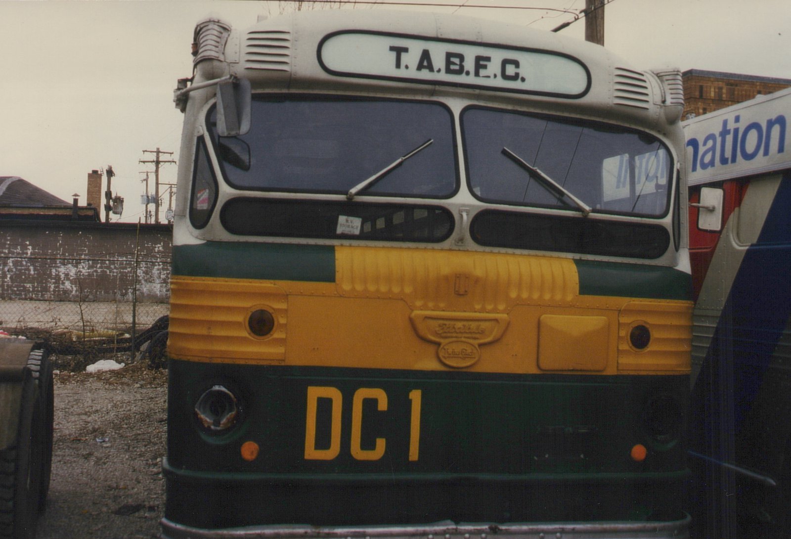 Front view of a green, yellow and white T.A.B.E.C. bus with the number DC 1 and a GM 'Schoolbus' emblem on its grille, parked in a gritty urban lot.