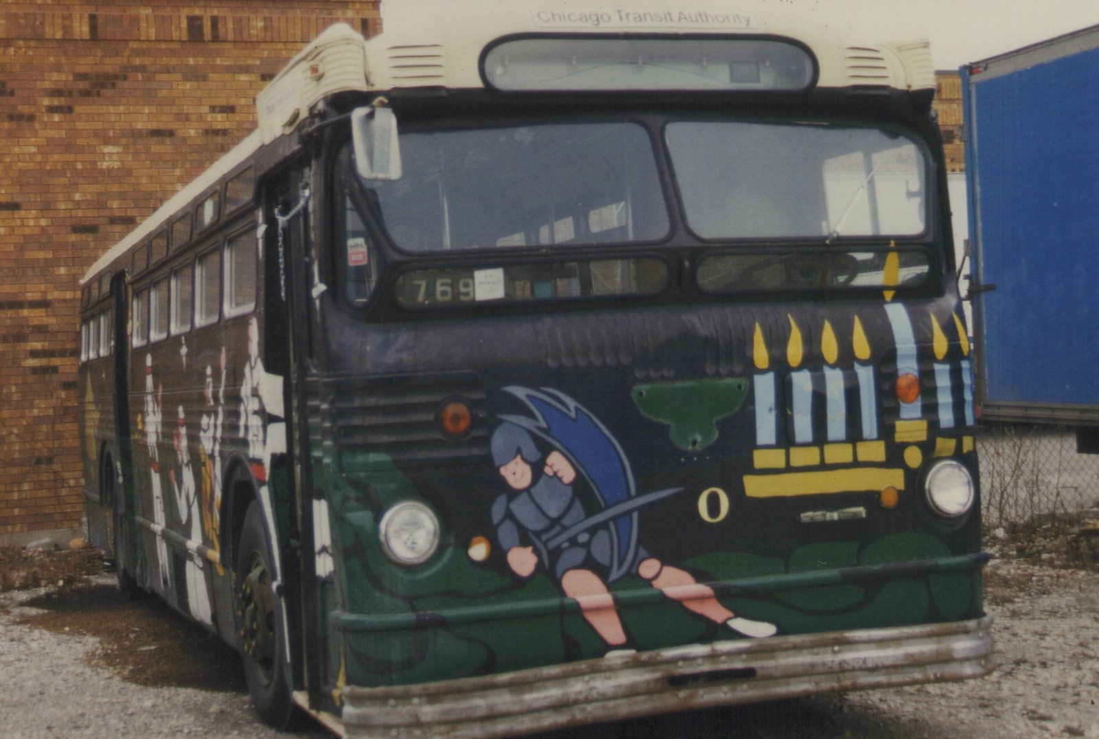 A dark green and black CTA bus with bus number 769 on the front, decorated with a colorful holiday mural featuring a cartoon knight and a menorah, parked next to a brick building on a gravel lot.