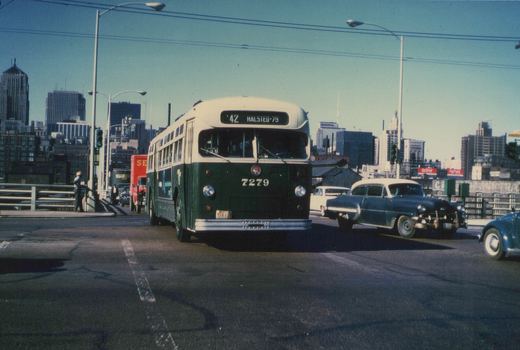 A green and cream CTA bus, number 7279, is stopped on a city bridge with a 1950s car beside it and skyscrapers in the background under a clear blue sky.