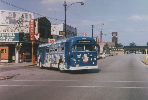 A blue Chicago Transit Authority bus with patriotic decals and 'Keep Chicago Clean' lettering is turning at a street corner, with storefronts and parked cars in the background.