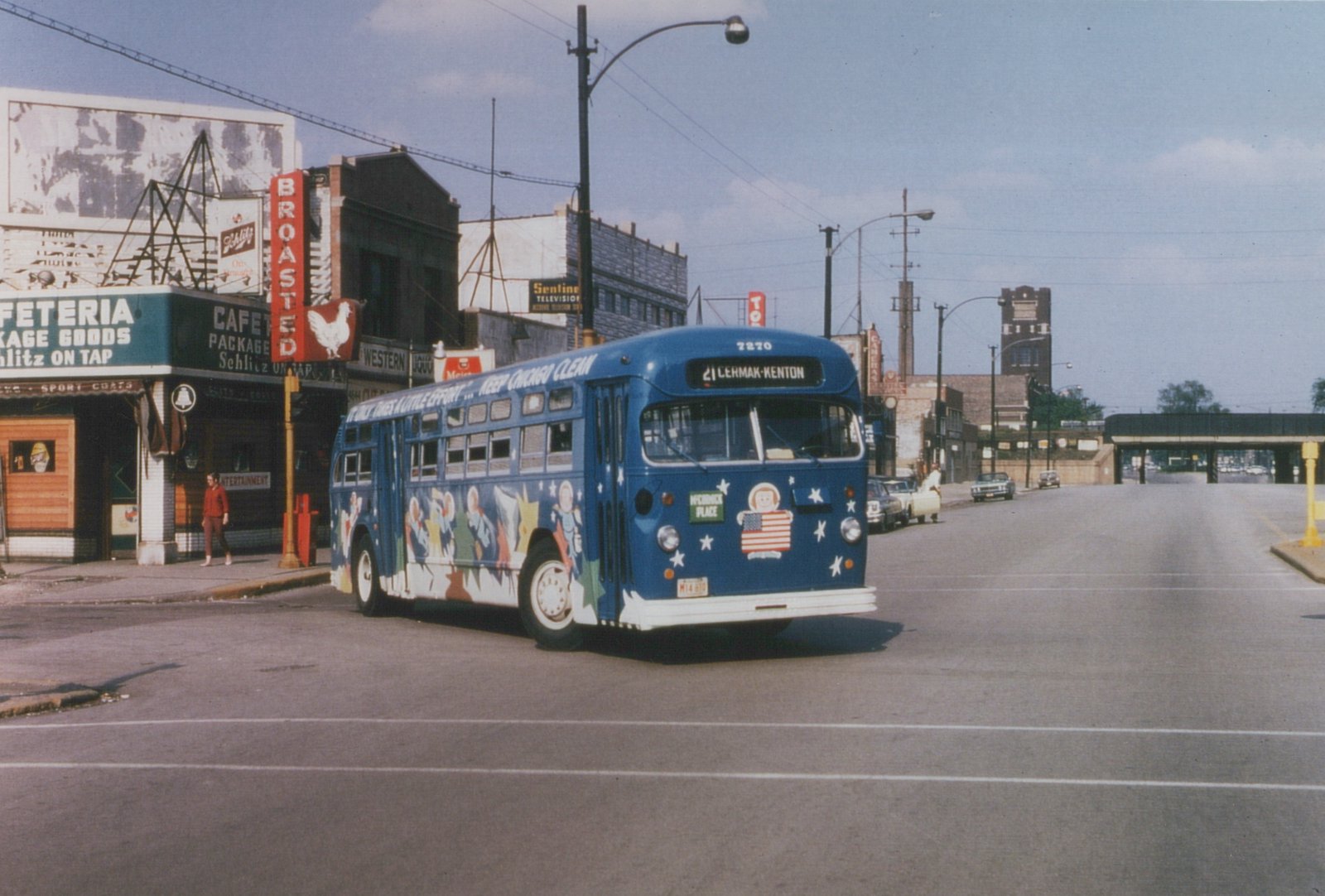 A blue Chicago Transit Authority bus with patriotic decals and 'Keep Chicago Clean' lettering is turning at a street corner, with storefronts and parked cars in the background.