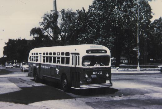 A black and white photo of a vintage city bus, number 6572, on a tree-lined street with several cars in the background.