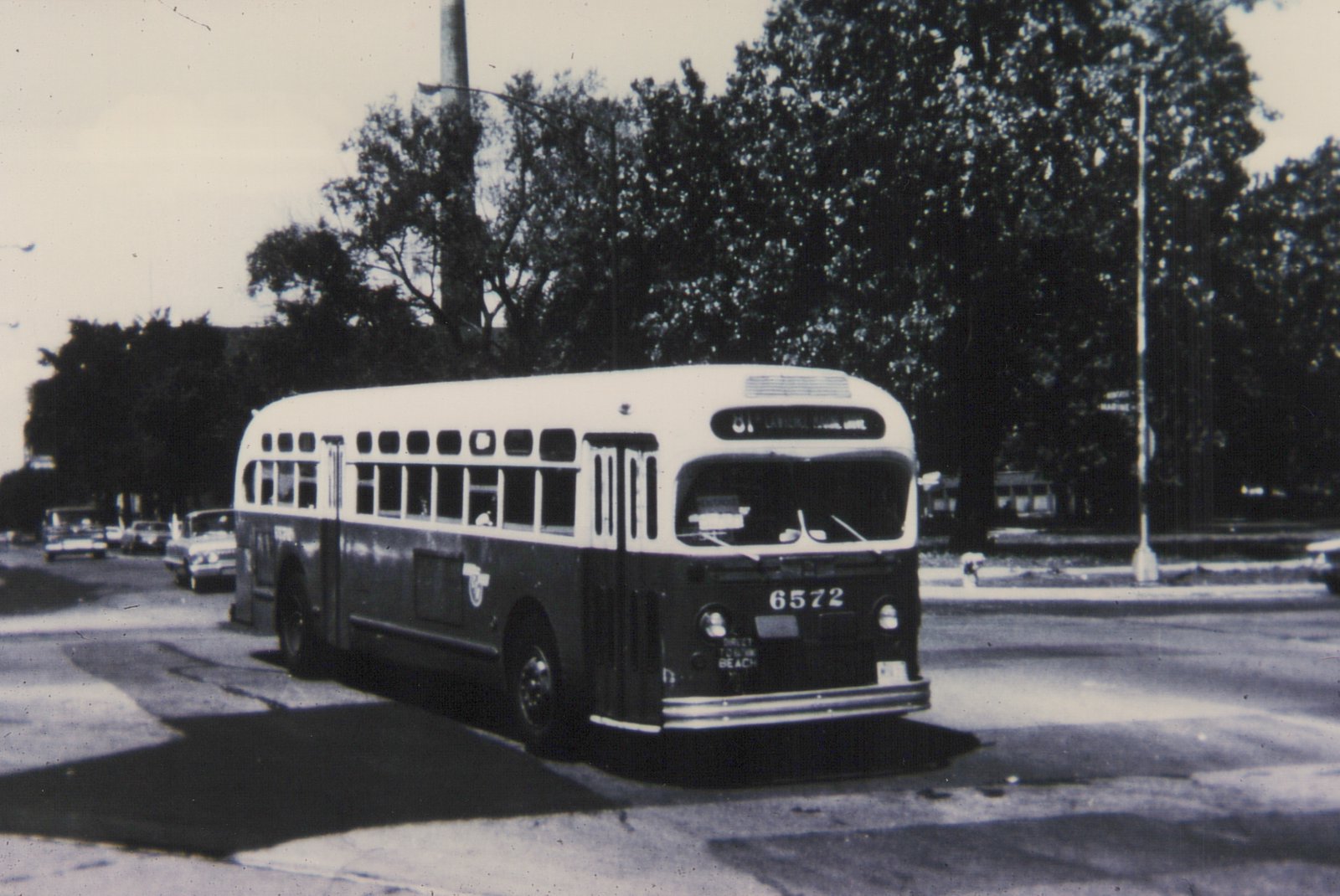 A black and white photo of a vintage city bus, number 6572, on a tree-lined street with several cars in the background.