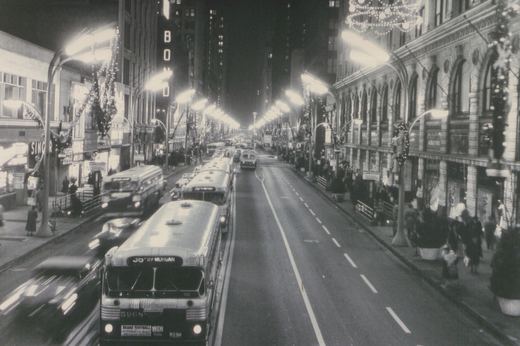 A nighttime street scene showing a line of Chicago Transit Authority buses on a busy, illuminated downtown street with Christmas decorations and pedestrians on the sidewalks.