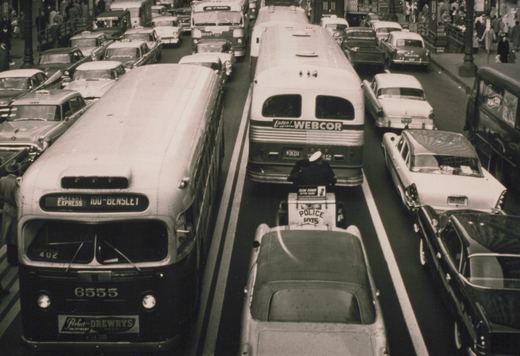 Two CTA buses, one showing route 100-Bensley and number 6555, are in heavy traffic with a police officer on a car behind them, amidst 1950s automobiles.
