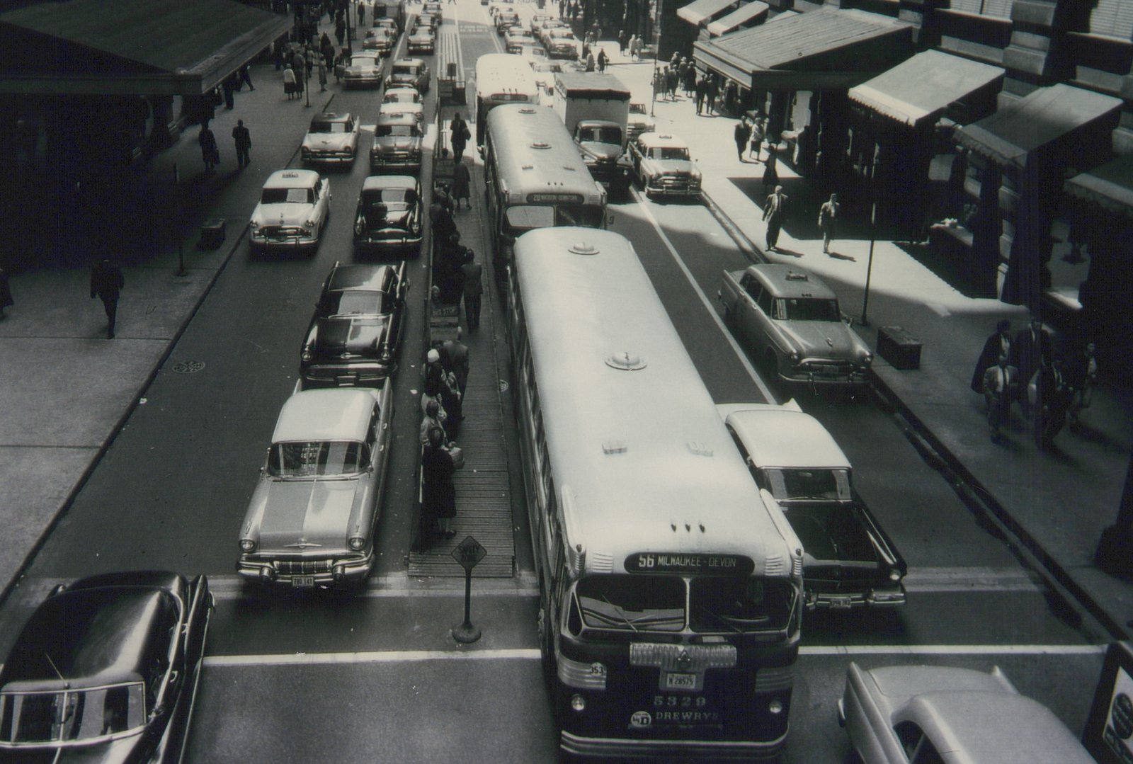 A black-and-white, high-angle shot of a busy Chicago street, showing a line of Drewry's buses and vintage cars at a bus stop.