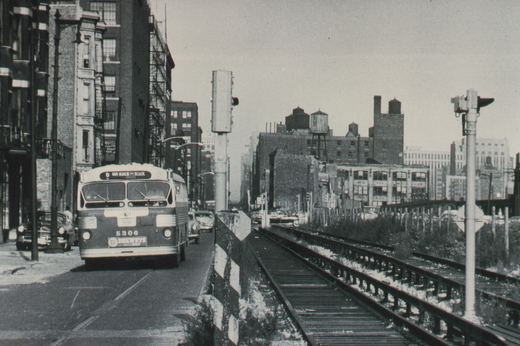 Front view of a dark-bodied CTA bus with the number 5306 and a 'Van Buren - Racine' destination sign, driving on a snowy city street next to elevated train tracks.