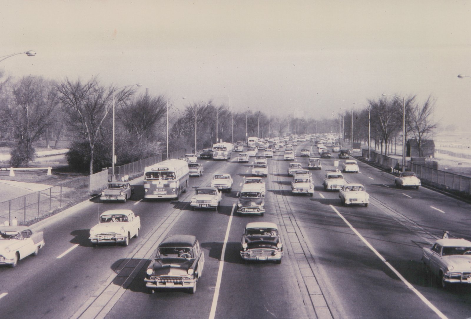 A black and white photo shows a Chicago Transit Authority bus driving on a wide highway with other cars; the surrounding trees are bare, indicating winter.