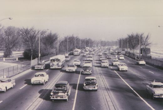 A black and white photo shows a Chicago Transit Authority bus driving on a wide highway with other cars; the surrounding trees are bare, indicating winter.