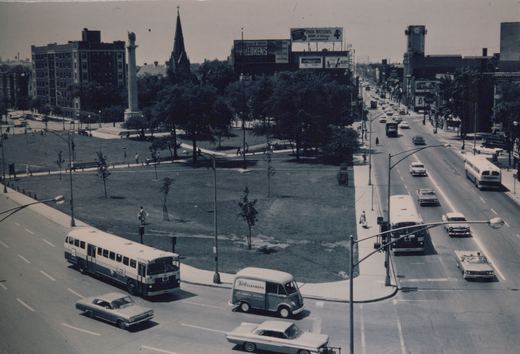 An elevated black and white photograph shows two Chicago Transit Authority buses on a wide city street next to a park with a tall monument, surrounded by 1960s cars and pedestrians.