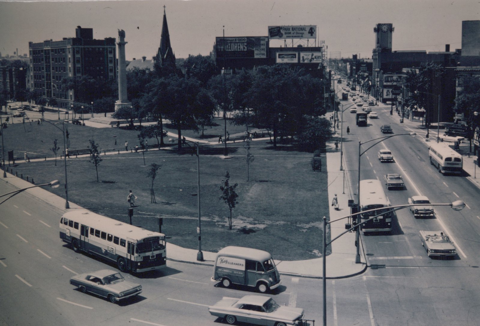 An elevated black and white photograph shows two Chicago Transit Authority buses on a wide city street next to a park with a tall monument, surrounded by 1960s cars and pedestrians.