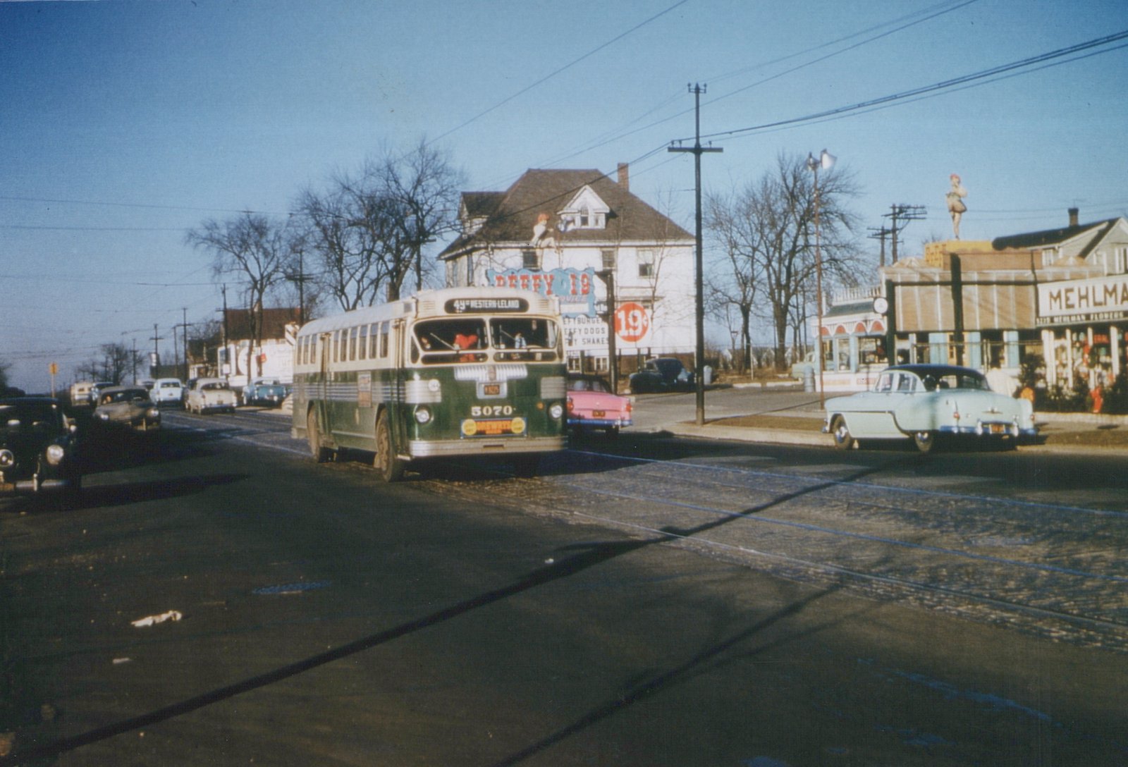 A green and cream Chicago Surface Lines bus, number 5070, drives down a street lined with 1950s cars, with store signs and bare trees in the background.