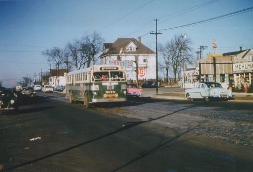 A green and cream Chicago Surface Lines bus, number 5070, drives down a street lined with 1950s cars, with store signs and bare trees in the background.