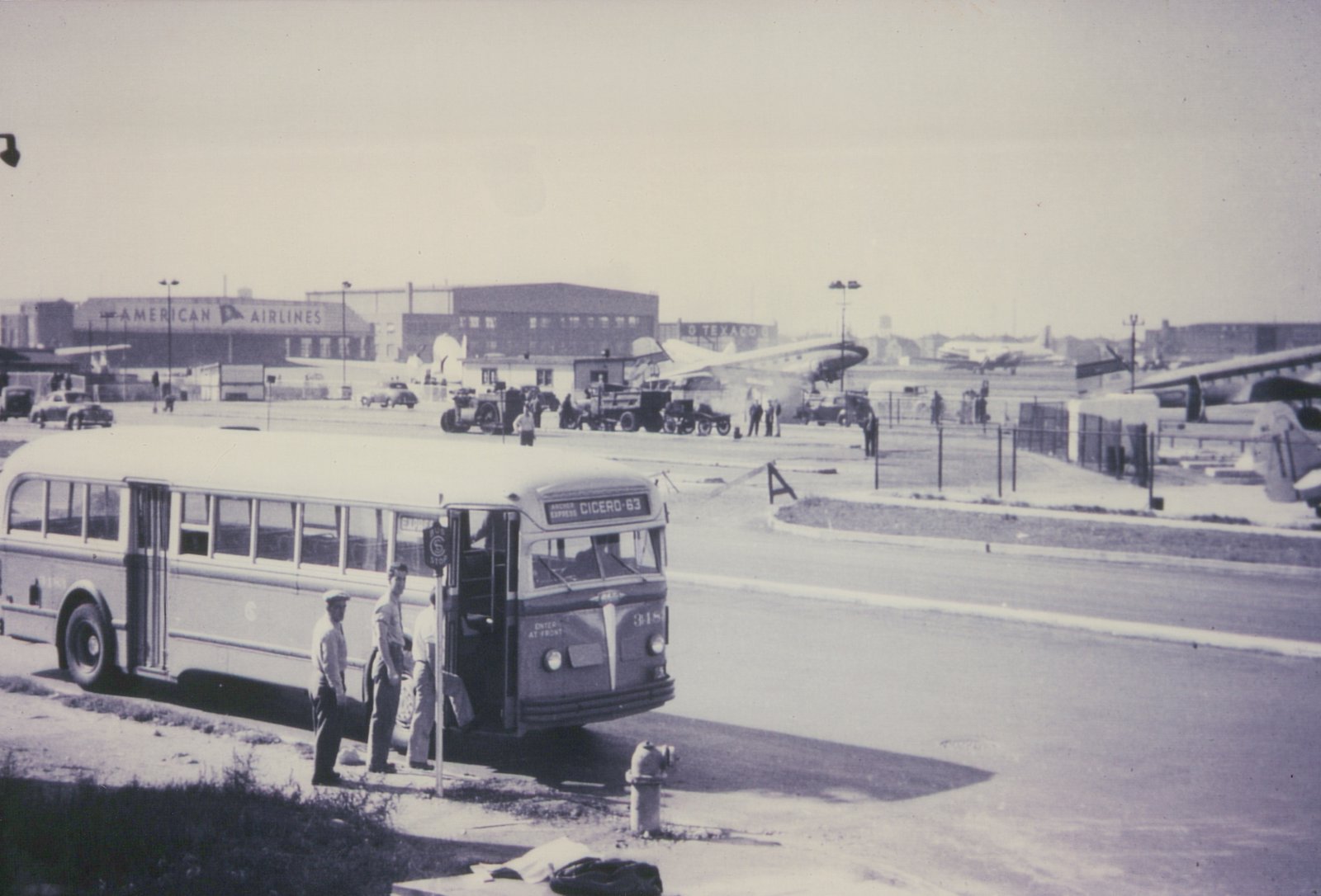 A 1940s motor bus from Chicago Surface Lines, fleet number 318, labeled 'Archer Express Cicero-63', is stopped at a bus stop on a sunny day, with American Airlines and Texaco buildings visible in the background.