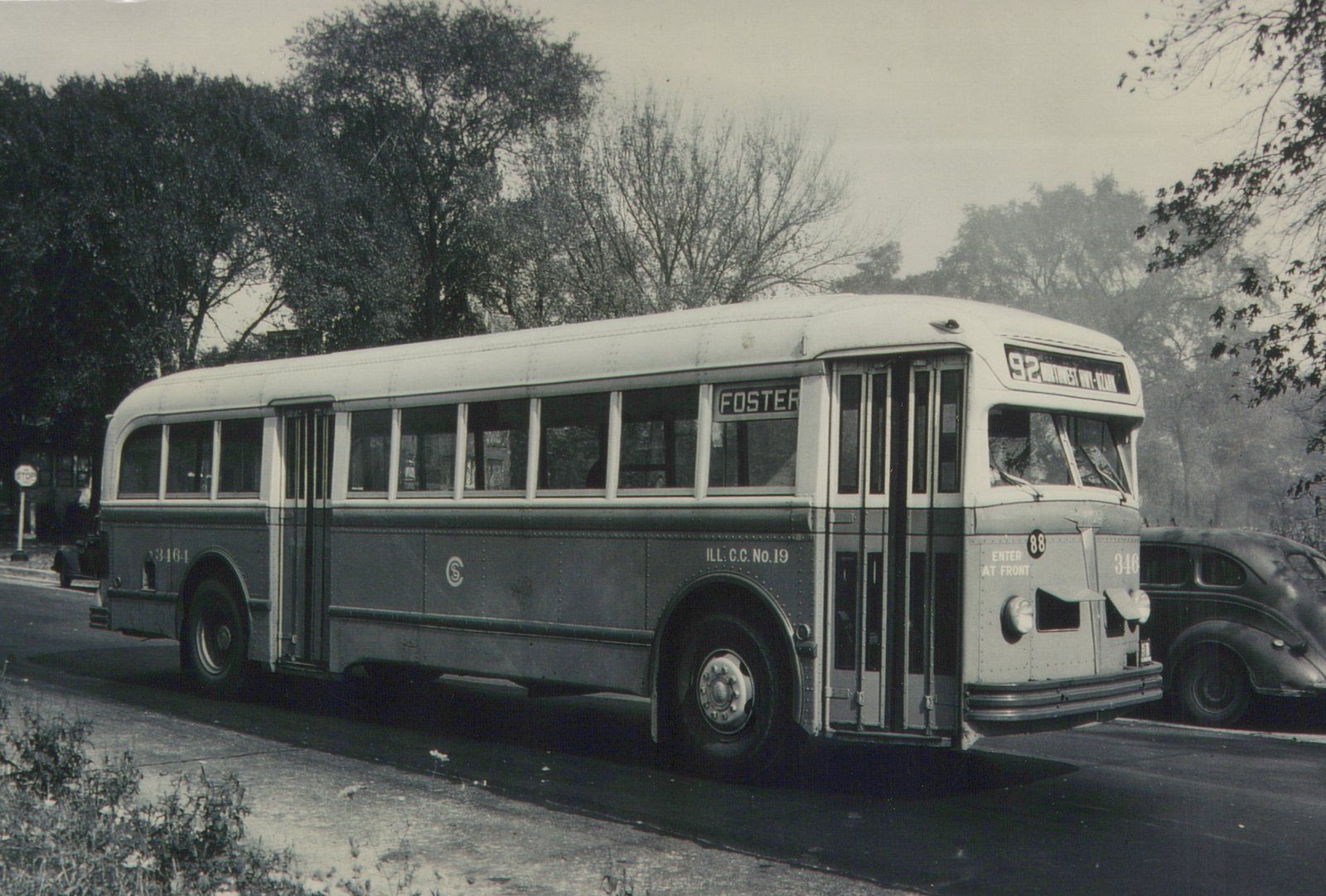A vintage black and white photo of a white-roofed motor bus with the number 346 and destination signs for route 92 on a street with trees and an old car in the background.