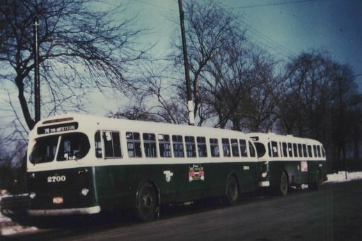 Two green and white Chicago Transit Authority buses from the 1940s or 1950s, numbered 2700, driving down a tree-lined street in winter, with snow on the ground.