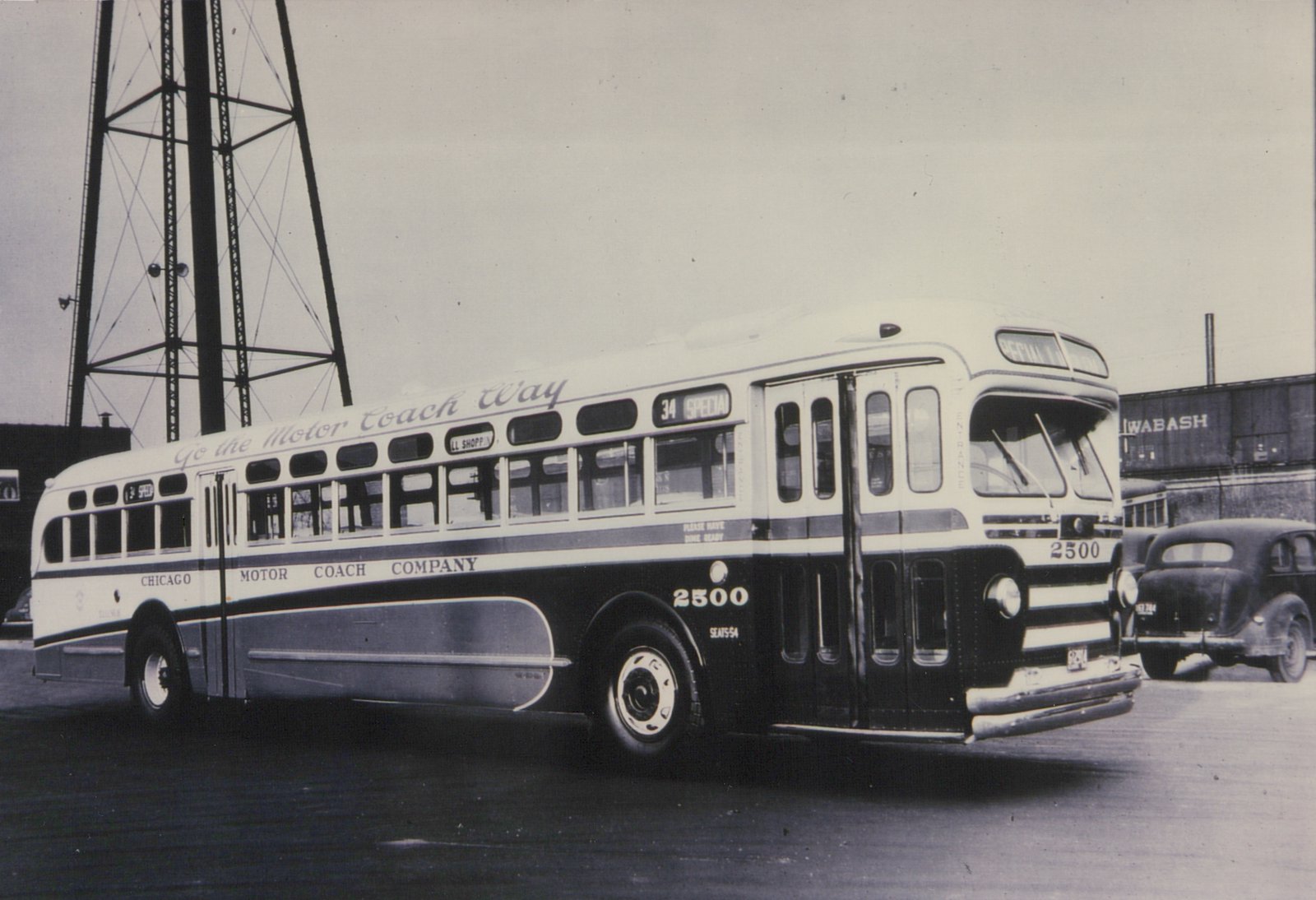A black and white photograph of a vintage Chicago Motor Coach Company bus, number 2500, parked on a street with a Wabash freight car and a classic car visible in the background.