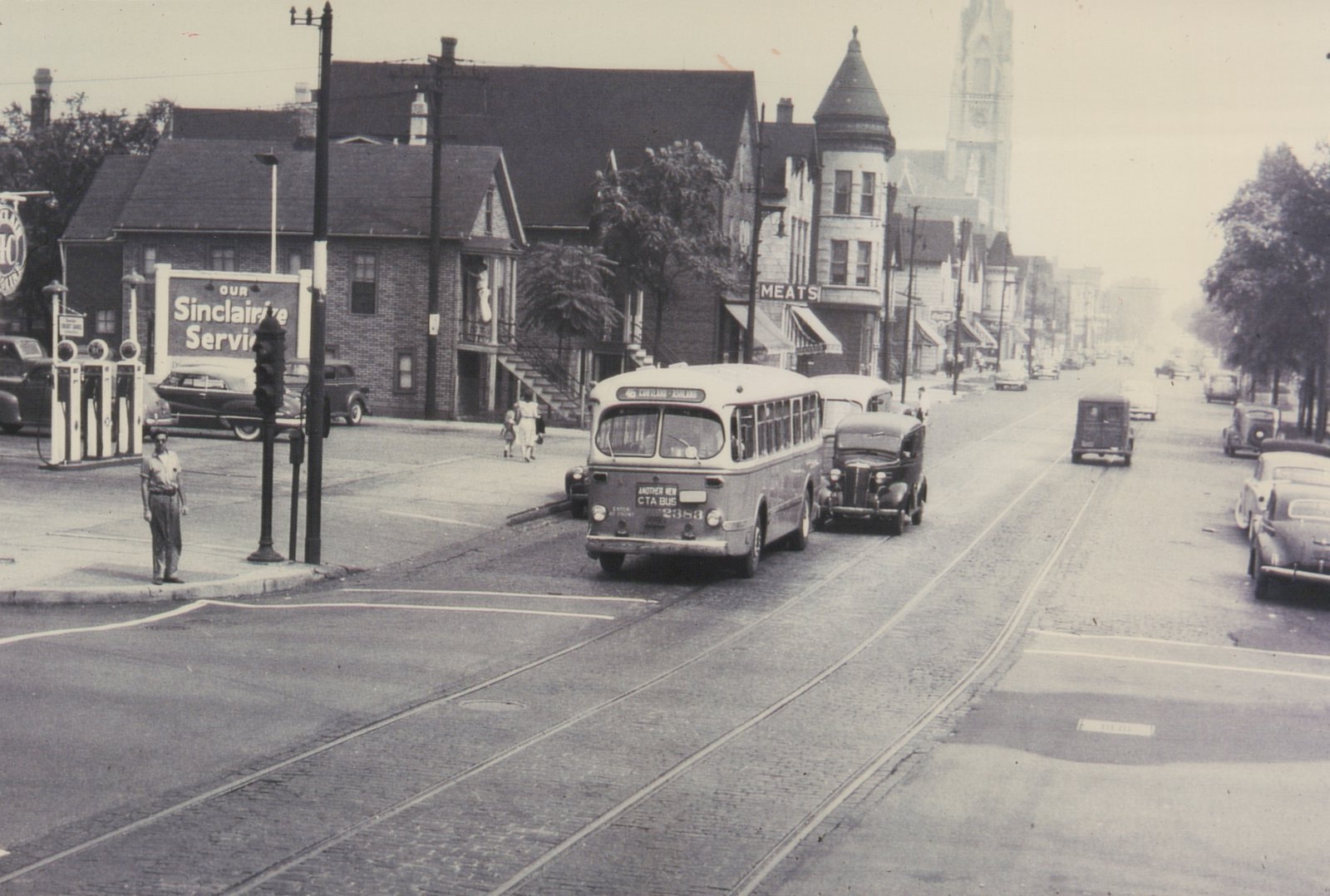 Rear view of a 1950s CTA bus on a city street with old cars, a Sinclair gas station, and a church steeple in the background.