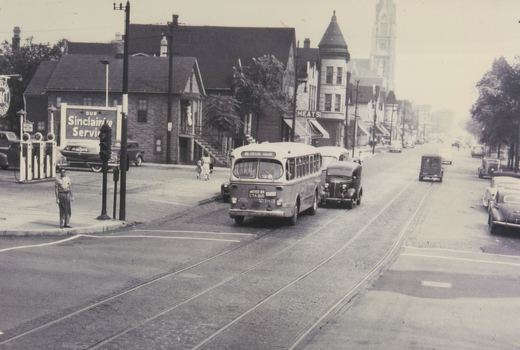 Rear view of a 1950s CTA bus on a city street with old cars, a Sinclair gas station, and a church steeple in the background.