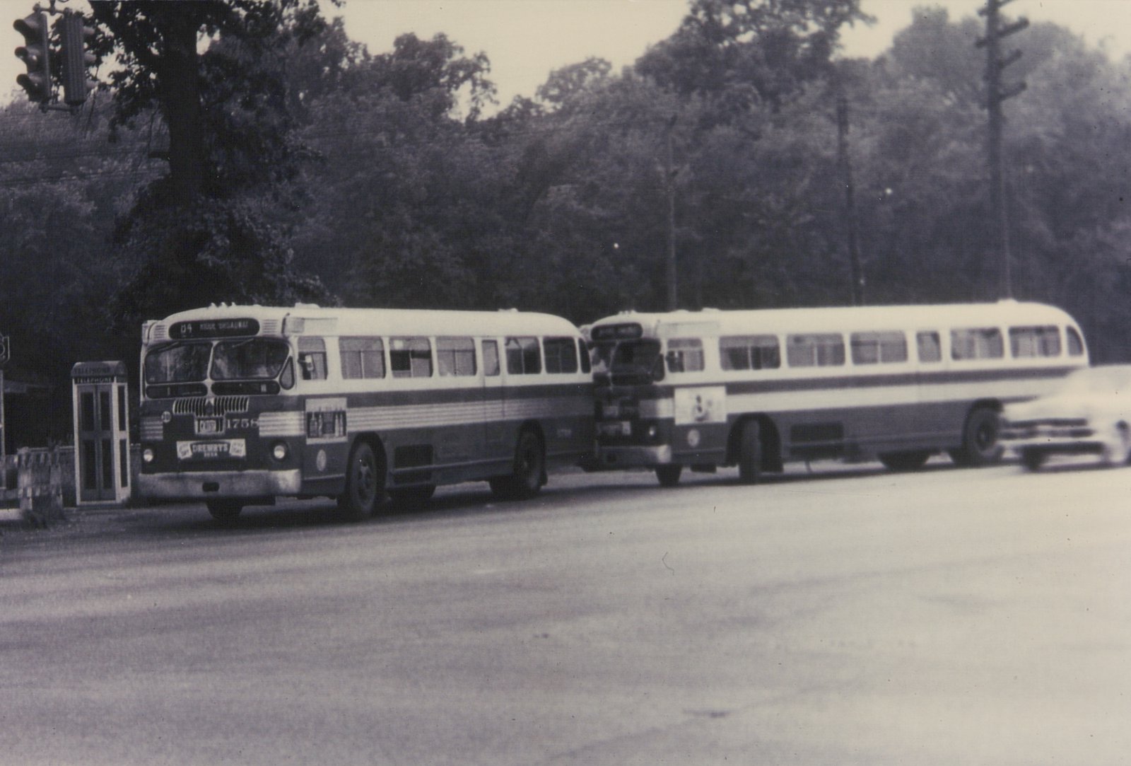 Two vintage city buses, one with the number 1756 on its front, are stopped on a tree-lined street with a telephone booth nearby.
