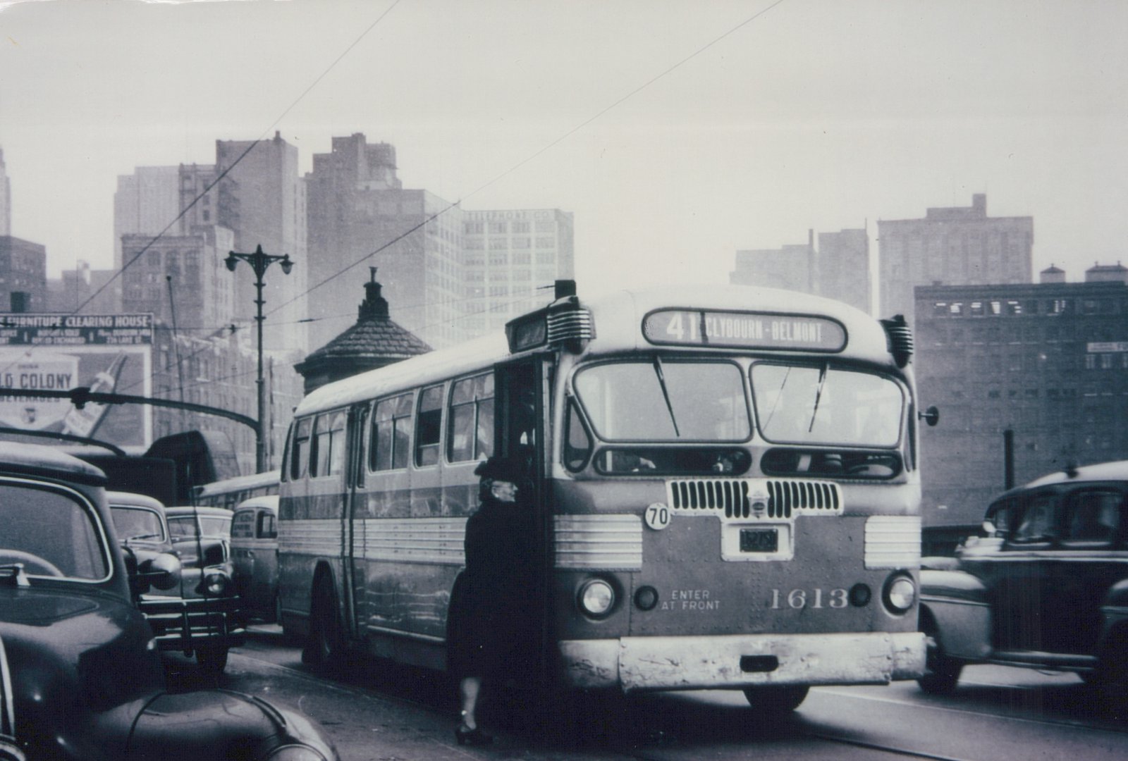 A black and white photo of a vintage motor bus on a city street with tall buildings in the background. A passenger is boarding, and other cars are visible.