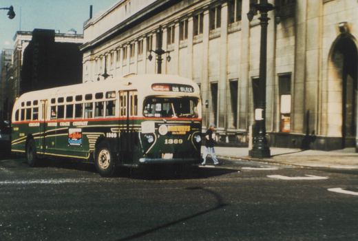 A green and cream Chicago Motor Coach Company bus numbered 1369 is on a city street, with a pedestrian crossing nearby and a large building in the background.