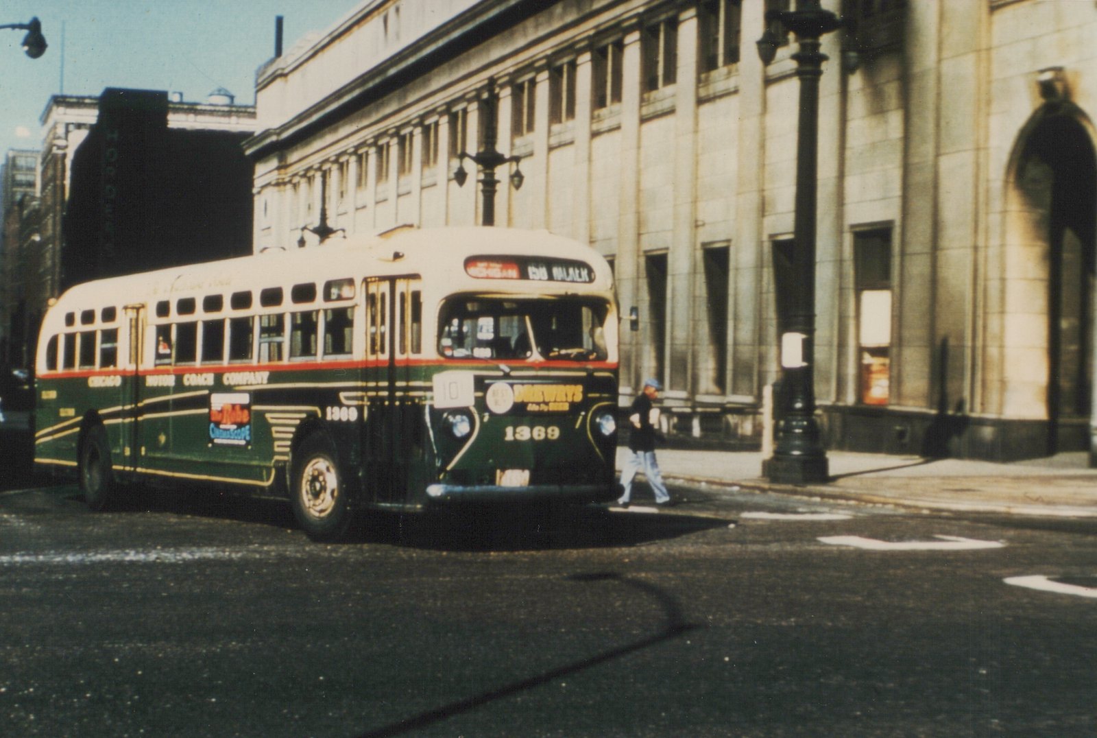 A green and cream Chicago Motor Coach Company bus numbered 1369 is on a city street, with a pedestrian crossing nearby and a large building in the background.