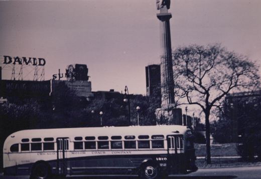 A black and white photo shows a side view of a white and dark-colored bus with 'CHICAGO MOTOR COACH COMPANY' lettered on its side, driving on a street in winter with bare trees and a tall column monument in the background.