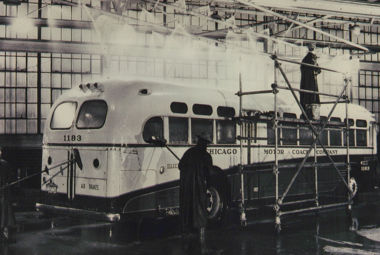 A black and white photo showing three workers washing a white and dark green bus with the number 1183 and 'Chicago Motor Coach Company' lettering inside a large, windowed garage.
