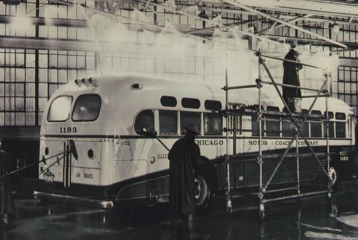 A black and white photo showing three workers washing a white and dark green bus with the number 1183 and 'Chicago Motor Coach Company' lettering inside a large, windowed garage.