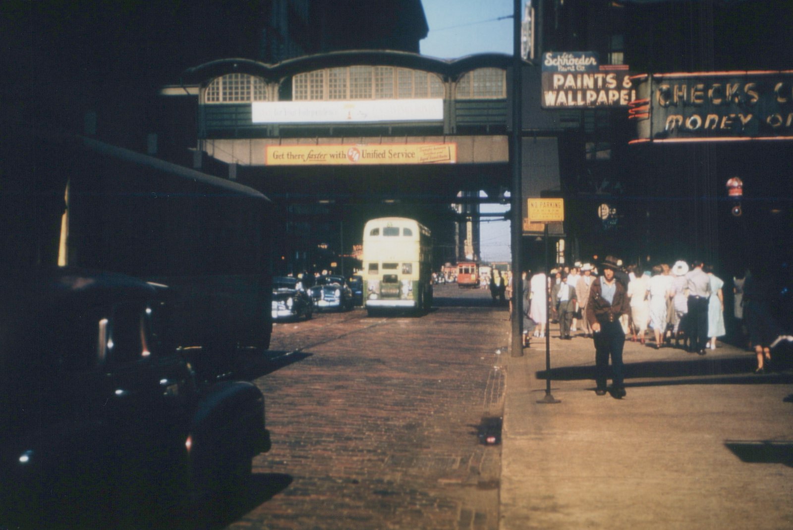 A street scene with a cream and green CTA bus and a red CTA streetcar under an elevated train structure, with pedestrians on the sidewalk and vintage cars parked alongside a brick-paved street.