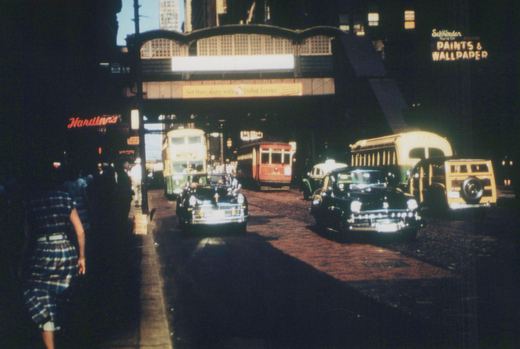 A street scene in downtown Chicago featuring a red streetcar, green and yellow buses, and vintage automobiles on a cobblestone street, with an elevated train structure overhead and neon signs on surrounding buildings.