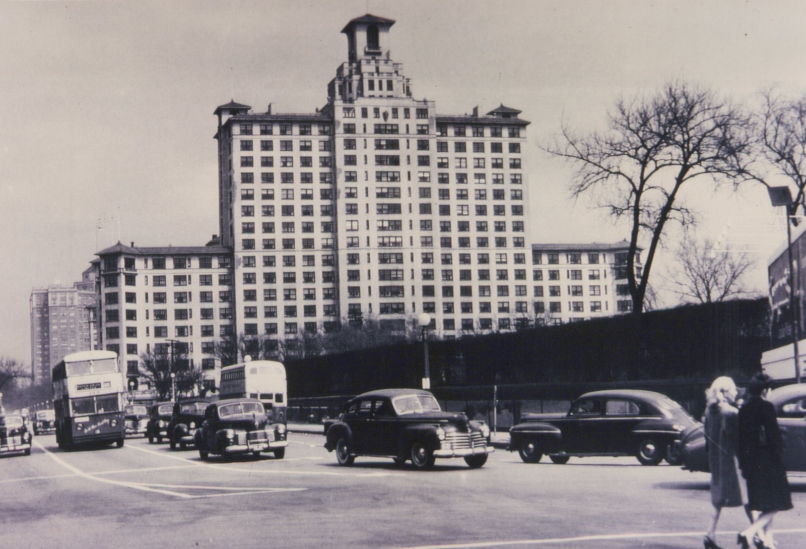 A black-and-white photograph of a city street in Chicago with double-decker buses, classic cars, and pedestrians, with a large apartment building in the background.
