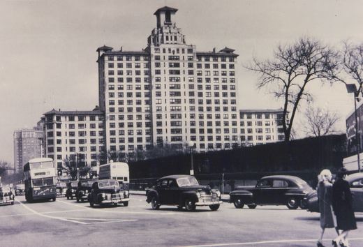 A black-and-white photograph of a city street in Chicago with double-decker buses, classic cars, and pedestrians, with a large apartment building in the background.