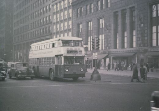 A black-and-white photo of a double-decker bus with 'Chicago Motor Coach Company' and '25 Jackson' on its signs, stopped at an intersection with pedestrians and 1940s cars nearby.