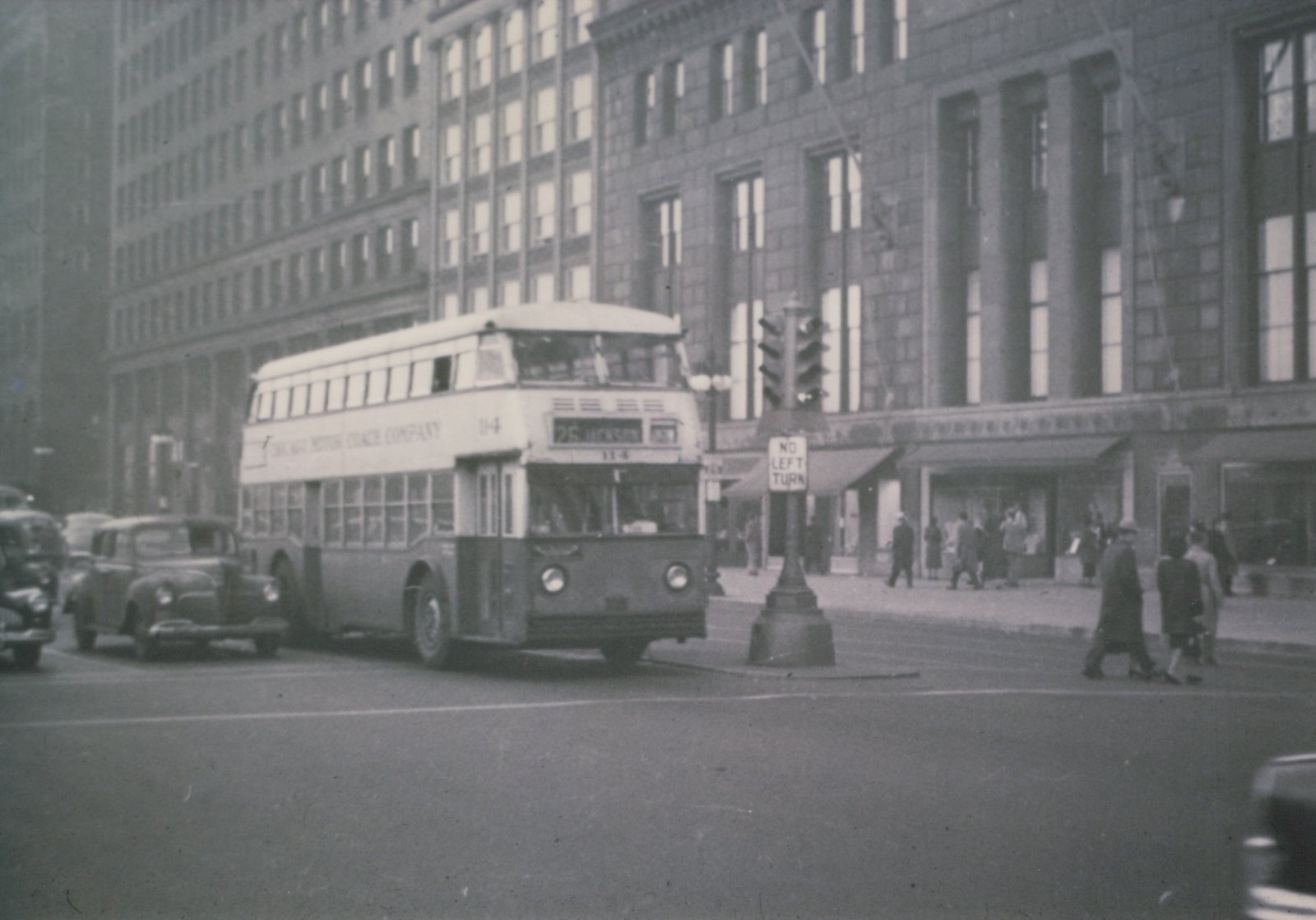 A black-and-white photo of a double-decker bus with 'Chicago Motor Coach Company' and '25 Jackson' on its signs, stopped at an intersection with pedestrians and 1940s cars nearby.