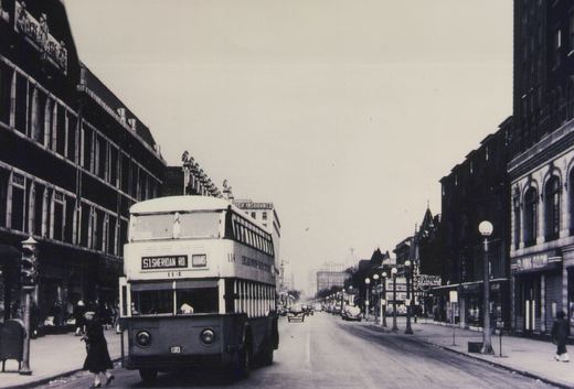 A black-and-white photo shows a vintage double-decker bus on a wide city street, with pedestrians on the sidewalks and multi-story buildings lining the road. The bus displays route 51 Sheridan Road and destination Adams.