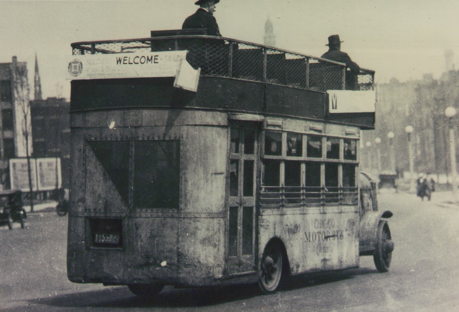 A black and white photograph of an old double-decker bus with 'CHICAGO MOTOR BUS' lettering on its side and the number 1155829 on its rear, driving down a street with two men on its open top deck.