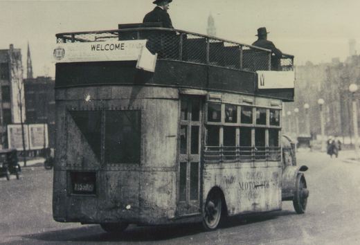 A black and white photograph of an old double-decker bus with 'CHICAGO MOTOR BUS' lettering on its side and the number 1155829 on its rear, driving down a street with two men on its open top deck.