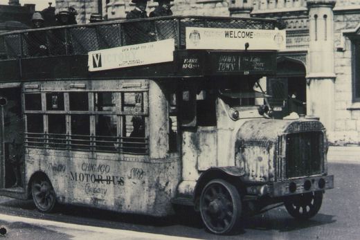 A vintage black and white photo of a double-decker bus labeled 'Chicago Motor Bus Company' with a 'Soldiers Welcome Sailors' sign on its roof, driving down a city street with passengers visible on the top deck.