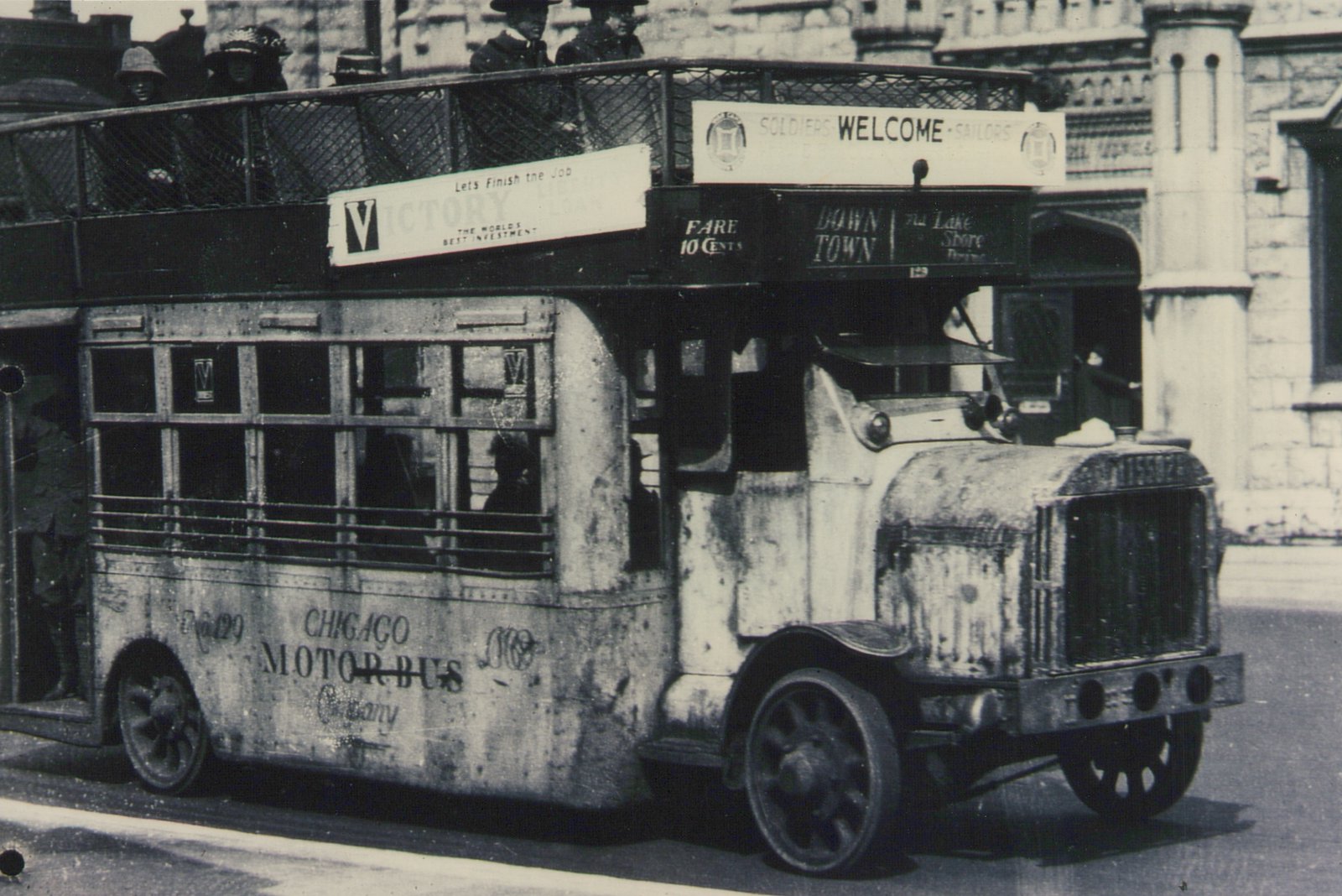 A vintage black and white photo of a double-decker bus labeled 'Chicago Motor Bus Company' with a 'Soldiers Welcome Sailors' sign on its roof, driving down a city street with passengers visible on the top deck.