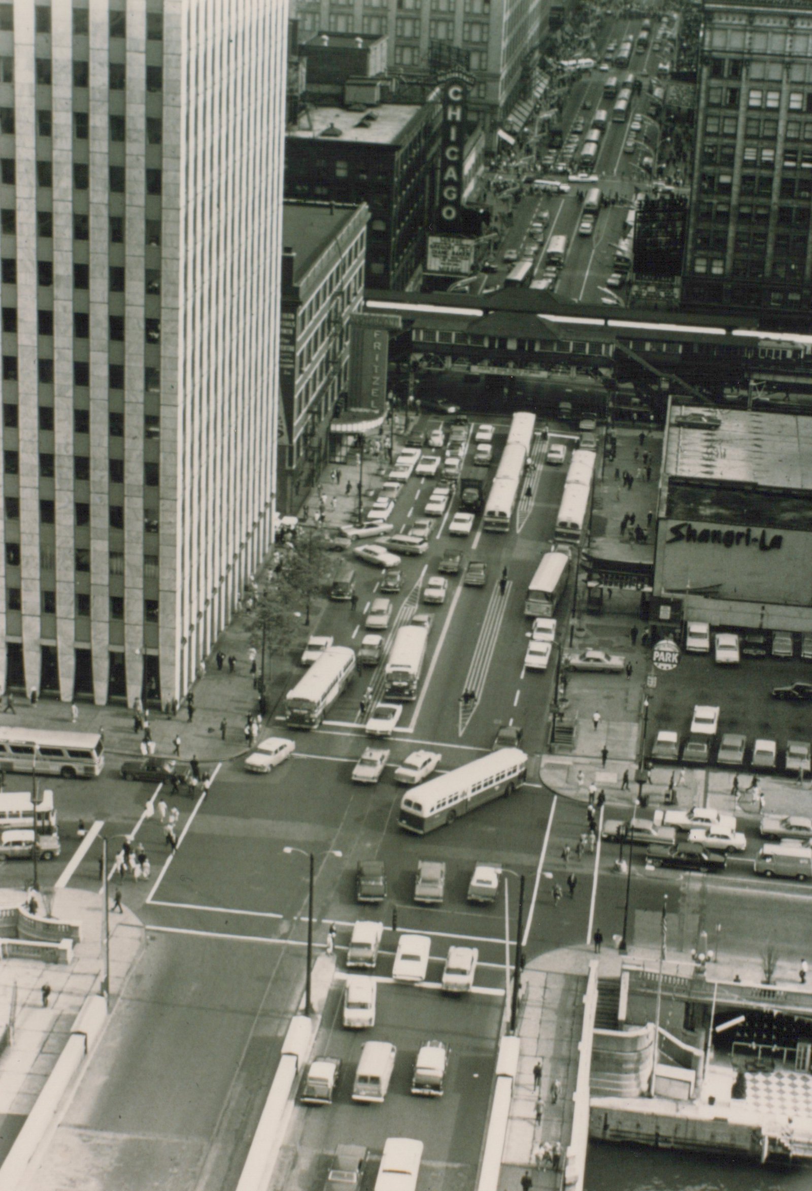 Black and white aerial photo of a bustling Chicago street corner in the 1960s, with many buses and cars, the Chicago Theater sign, and surrounding buildings.