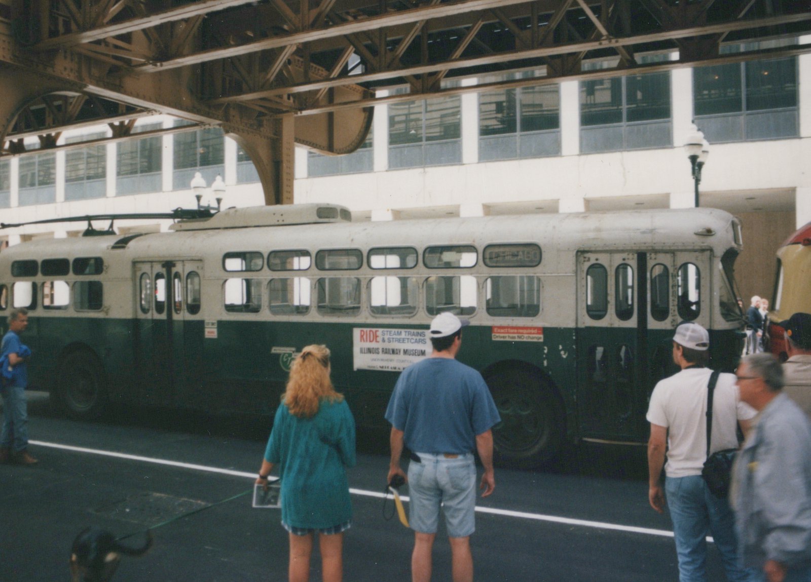 A vintage Chicago trolleybus with a cream and green livery, showing a 'CHICAGO' destination sign, parked under an elevated rail structure with several people looking at it.