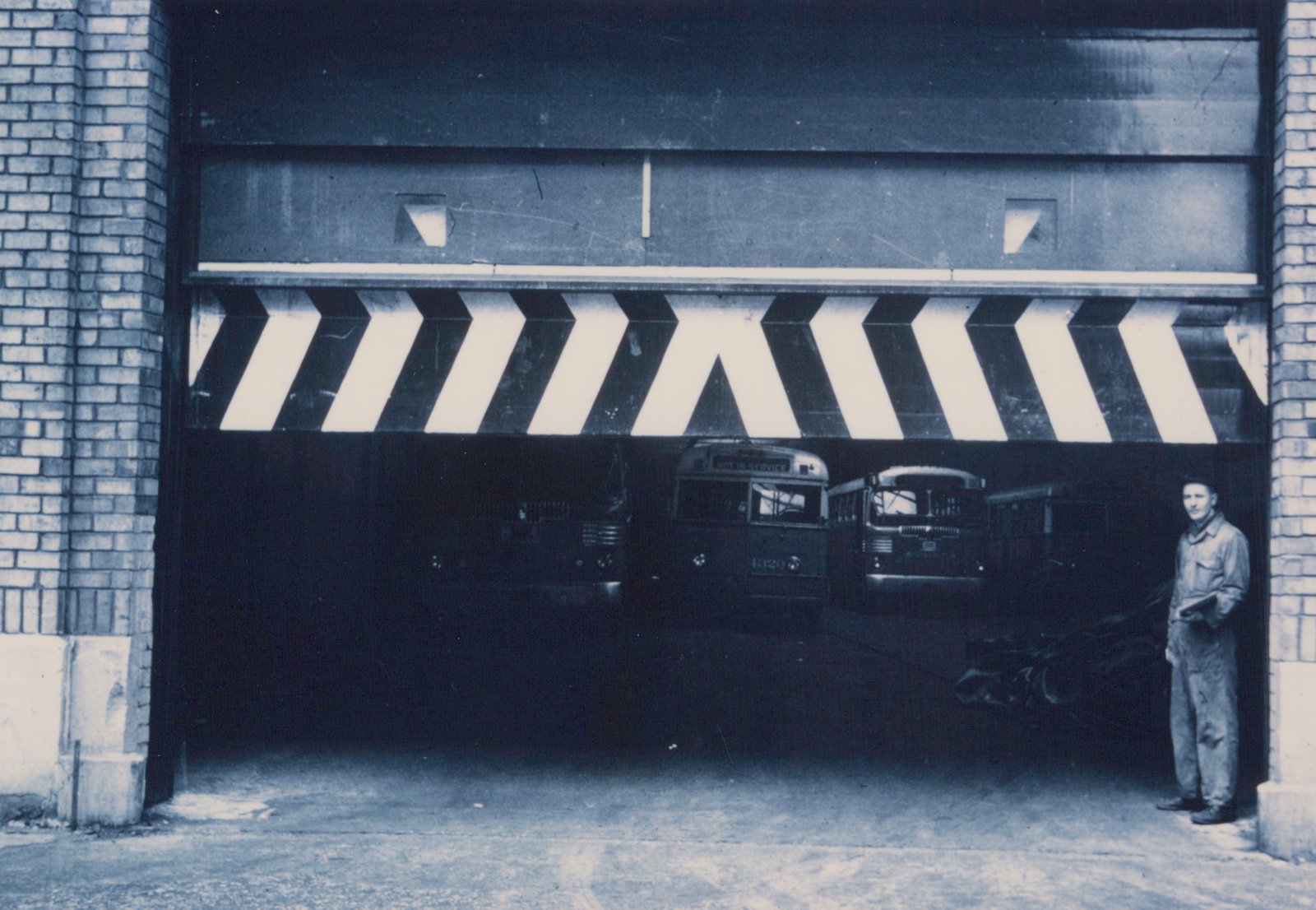 A black and white photo showing a line of dark-colored buses inside a garage with striped warning markings on the door; one bus has the number 1320 and a 'NOT IN SERVICE' sign; a man in overalls stands to the right.